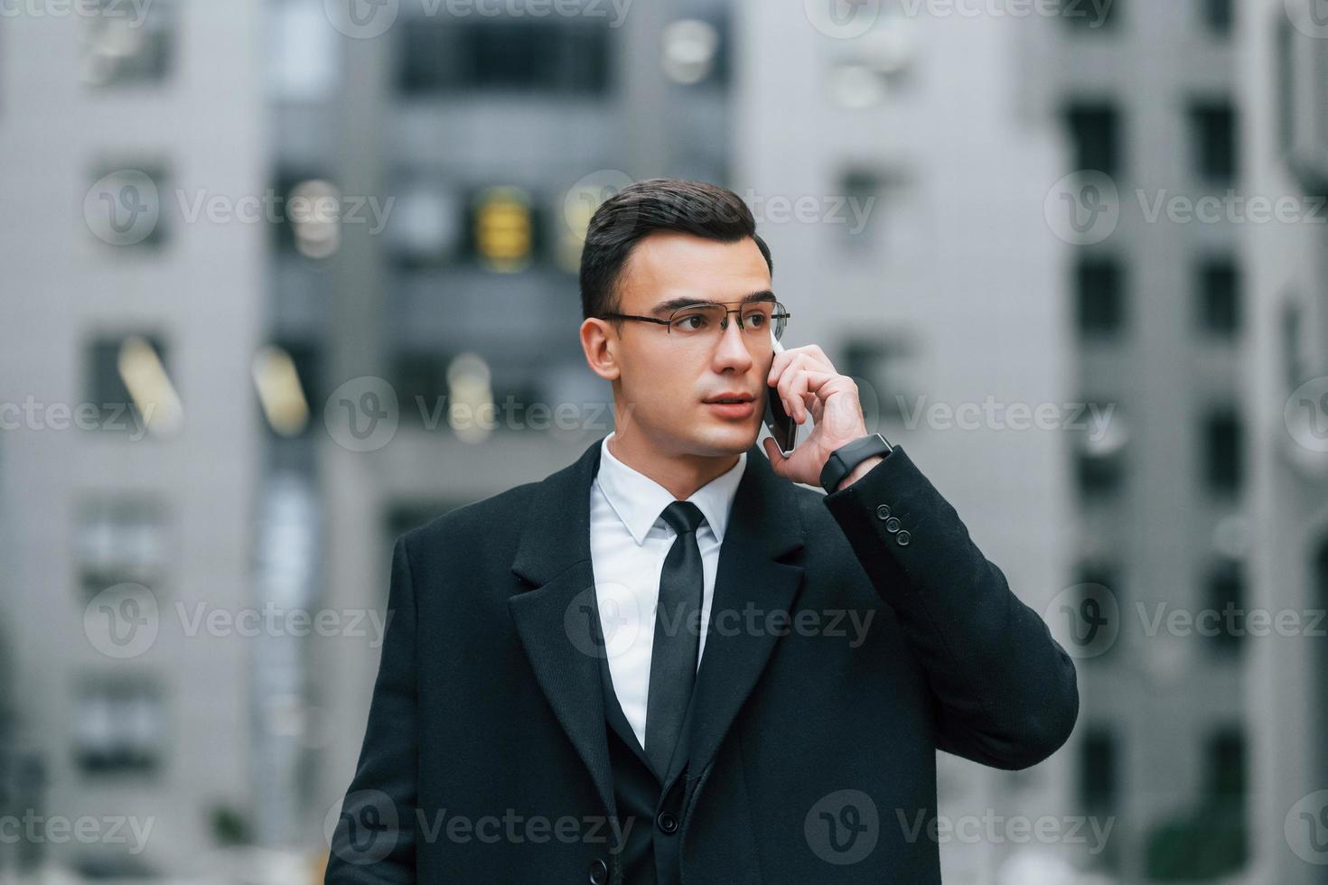 Talking by phone. Businessman in black suit and tie is outdoors in the city photo