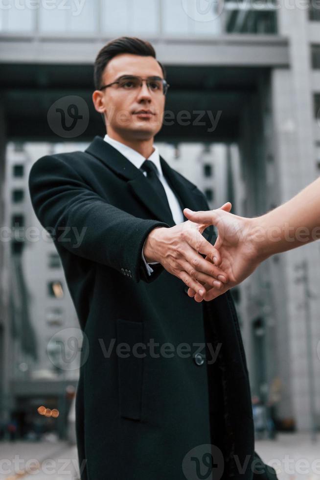 Doing handshake. Businessman in black suit and tie is outdoors in the city photo