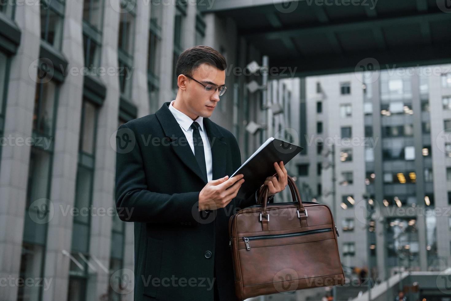 With notepad in hands. Businessman in black suit and tie is outdoors in the city photo
