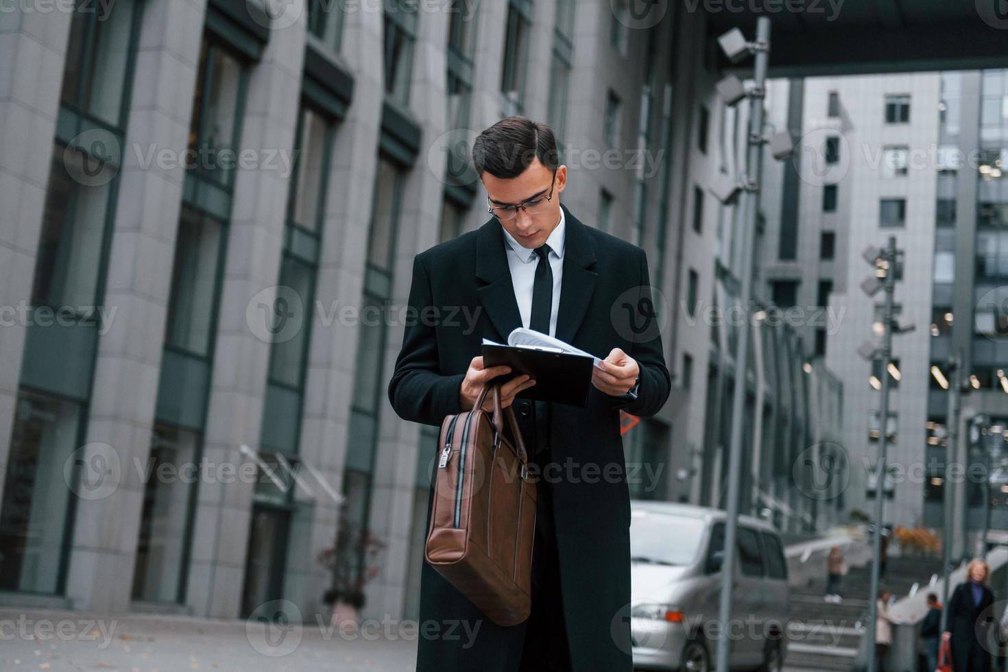 With notepad in hands. Businessman in black suit and tie is outdoors in the city photo