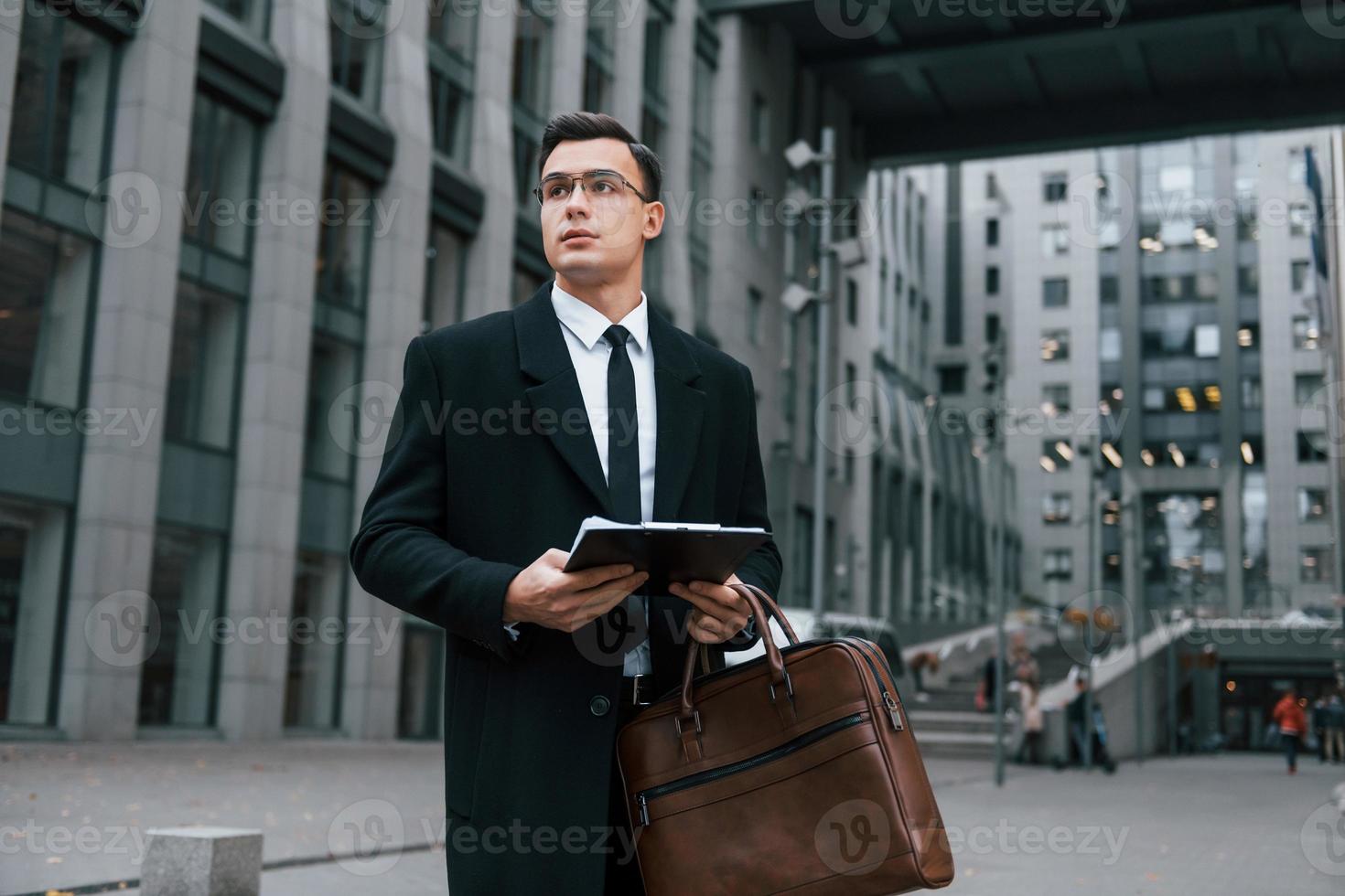 With notepad in hands. Businessman in black suit and tie is outdoors in the city photo