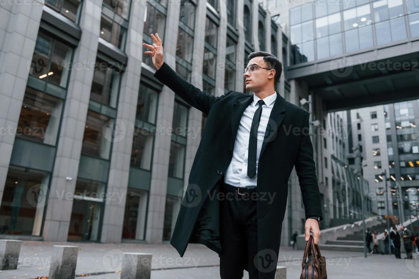 levanta su mano derecha. hombre de negocios con traje negro y corbata está al aire libre en la ciudad foto