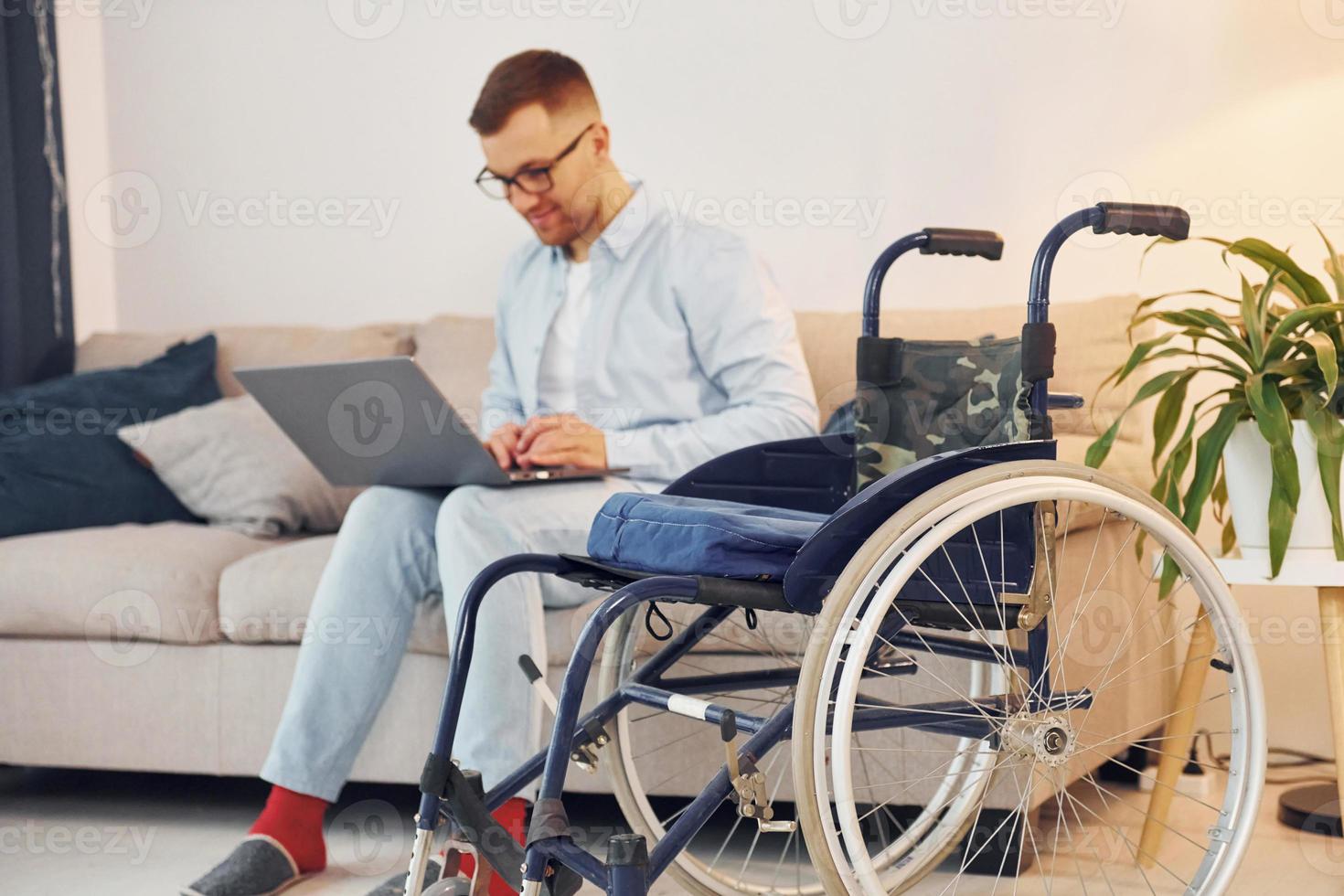 Sitting on sofa with laptop on legs. Disabled man in wheelchair is at home photo