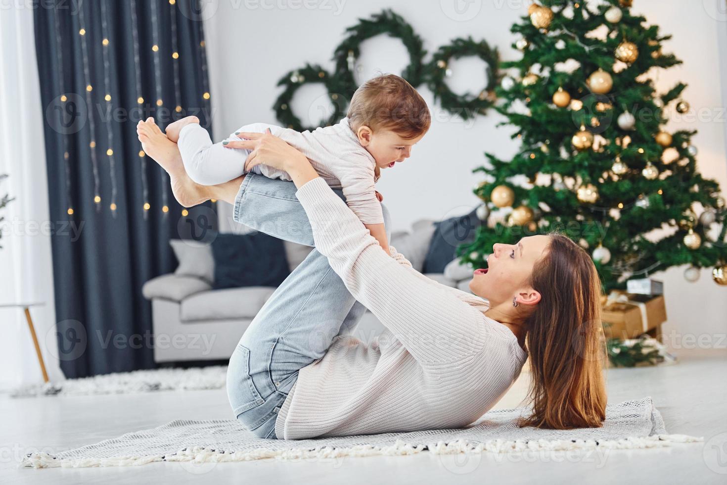 Laying down on the ground and having fun. Mother with her little daughter is indoors at home together photo