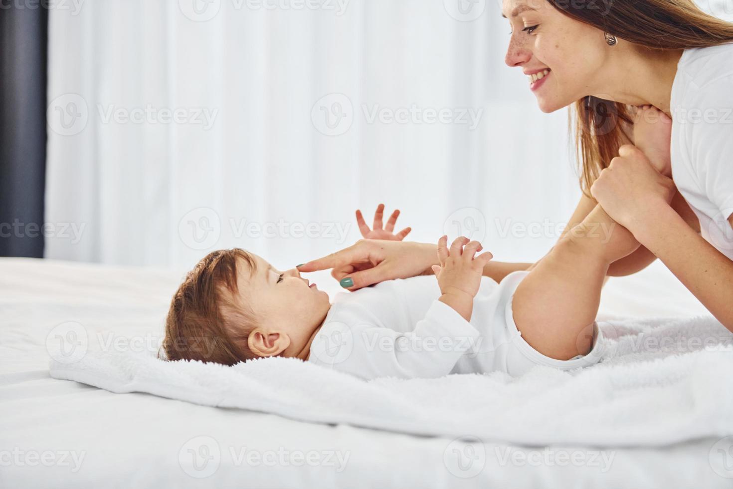 On the white bed. Mother with her little daughter is indoors at home together photo