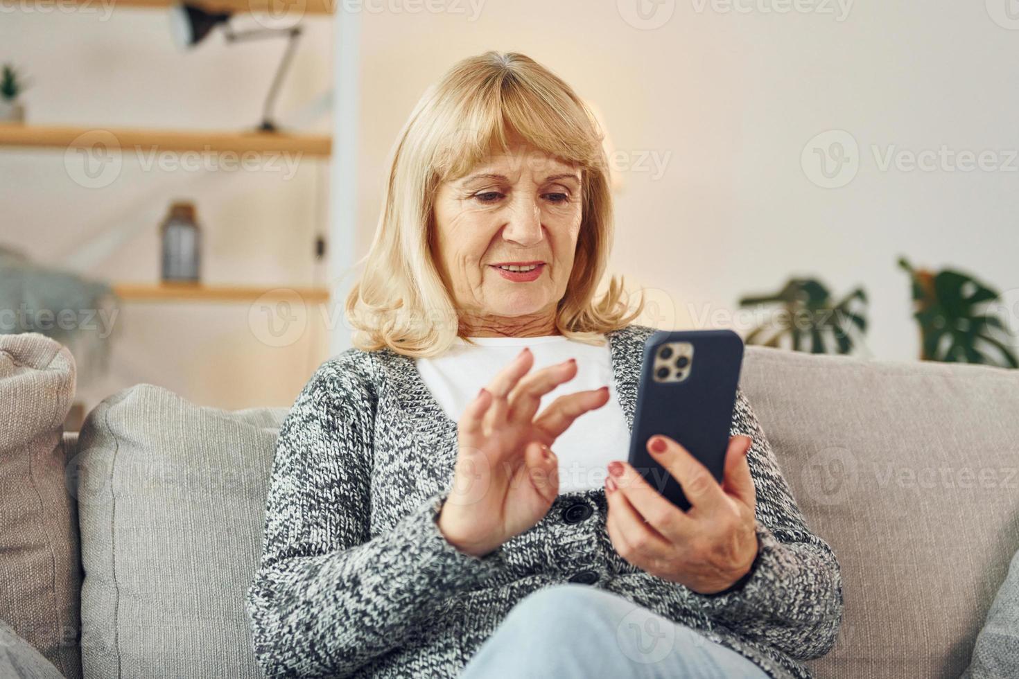 sentado y sosteniendo el teléfono. mujer mayor con cabello rubio está en casa foto