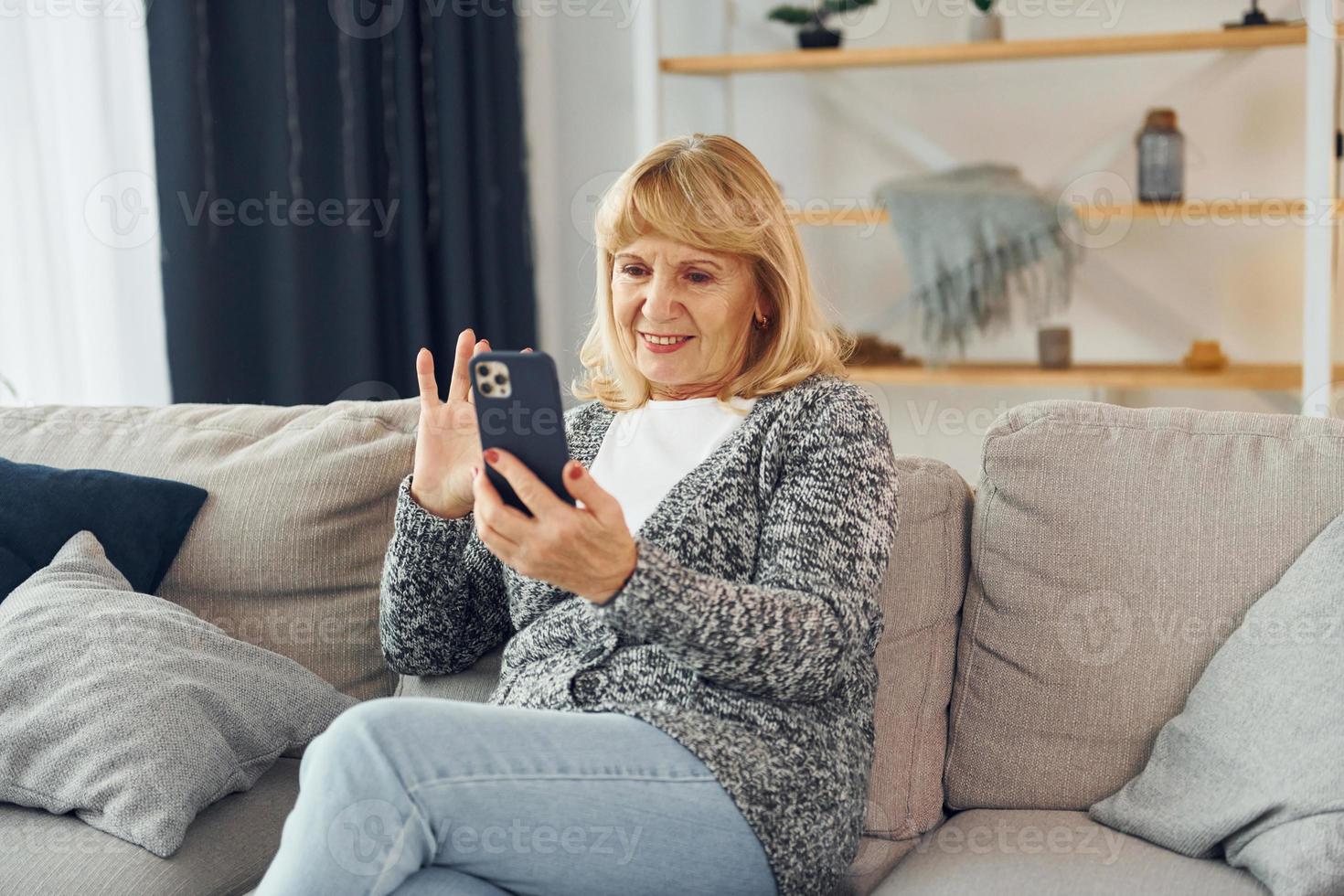 Sitting and holding phone. Senior woman with blonde hair is at home photo