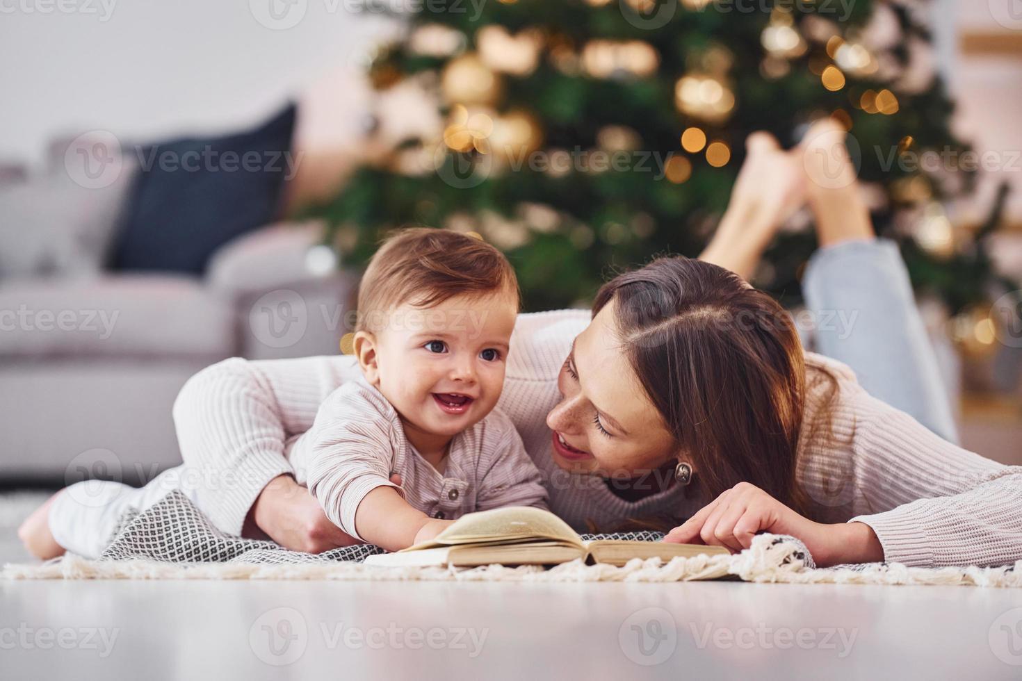 With book that is on the ground. Mother with her little daughter is indoors at home together photo