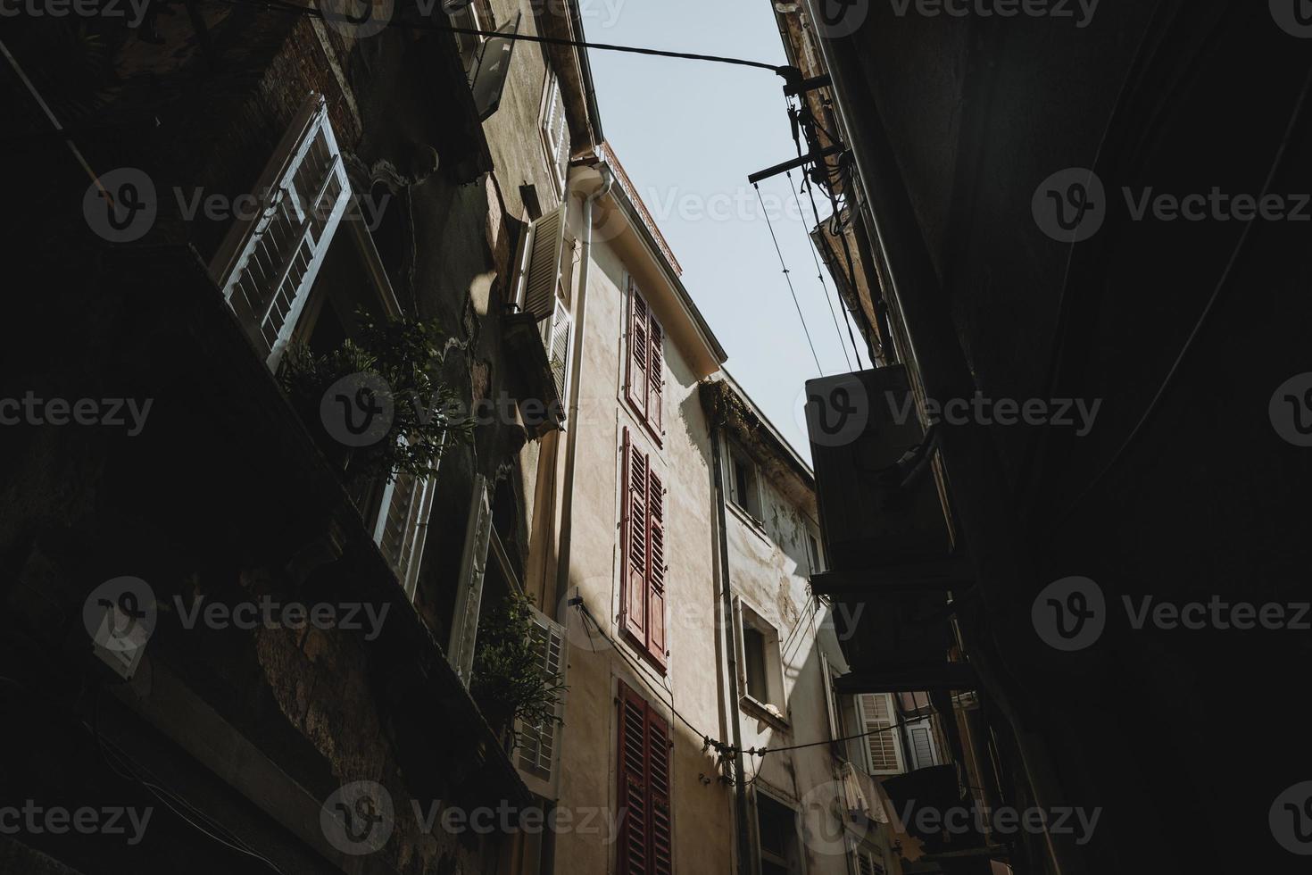 Bottom-up view to the tops of old buildings enlightened by the summer sun from the shadow of the street in historical town of Piran in Slovenia with the clear blue sky above photo