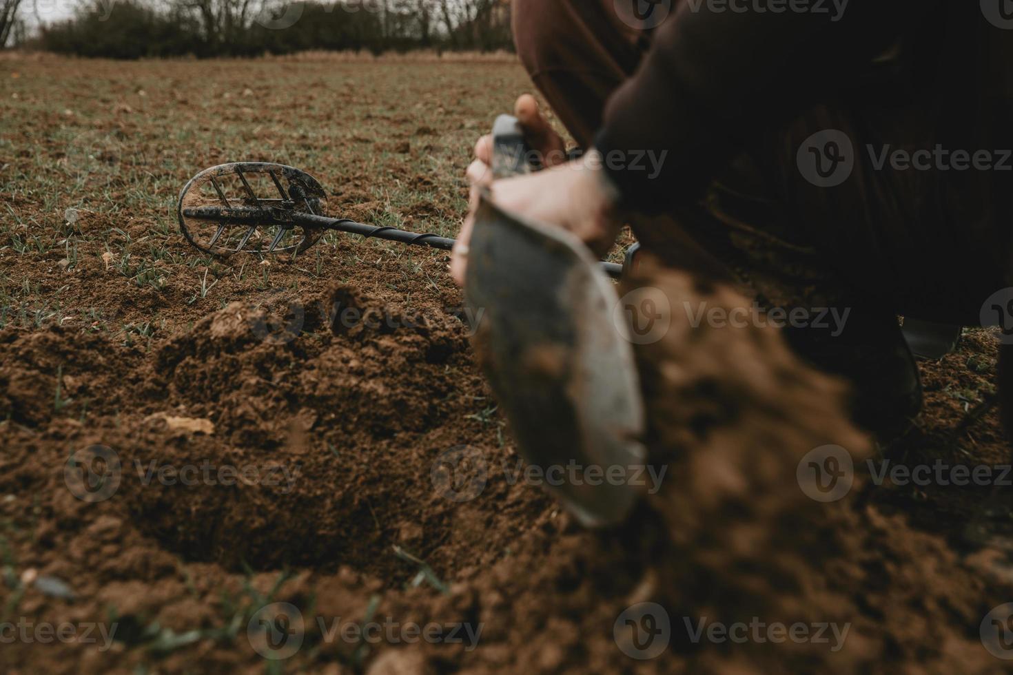 detalle de las manos del hombre con pala, durante la excavación de un agujero en el suelo después de la señal positiva del detector de metales con detector de metales acostado en segundo plano foto