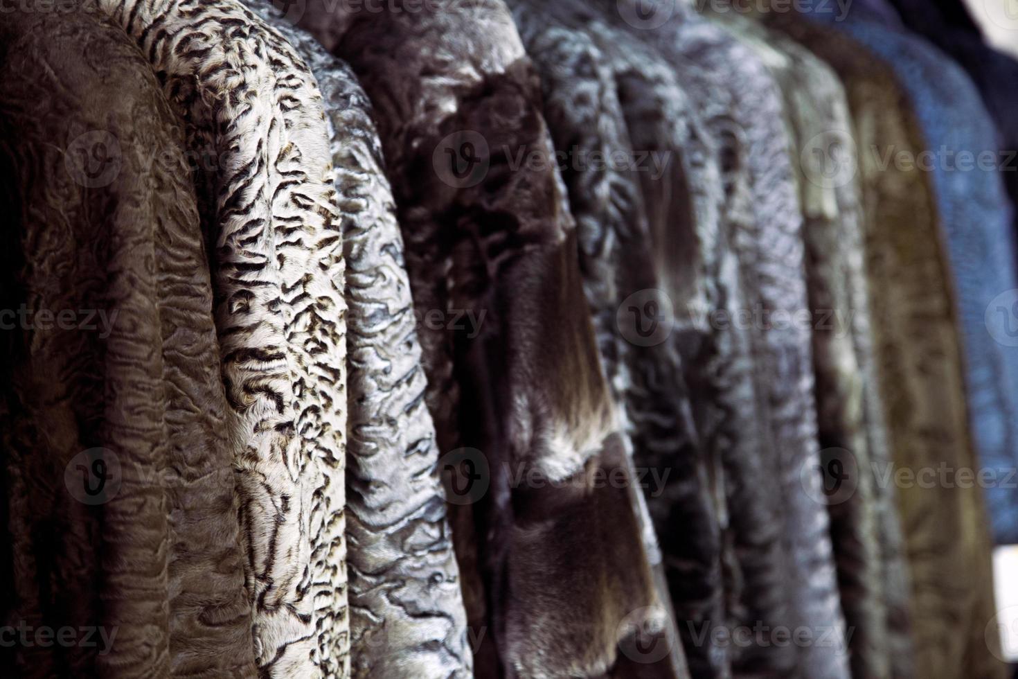 A close up of a fur coat from a natural fur of astrakhan of different colors  brown, gray and black, hang in one row in the store photo