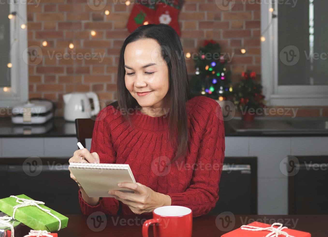 Asian woman wearing red knitted sweater sitting  at table with red cup of coffee and gift boxes  in the kitchen with Christmas decoration, using pen writing name list on notebook. photo
