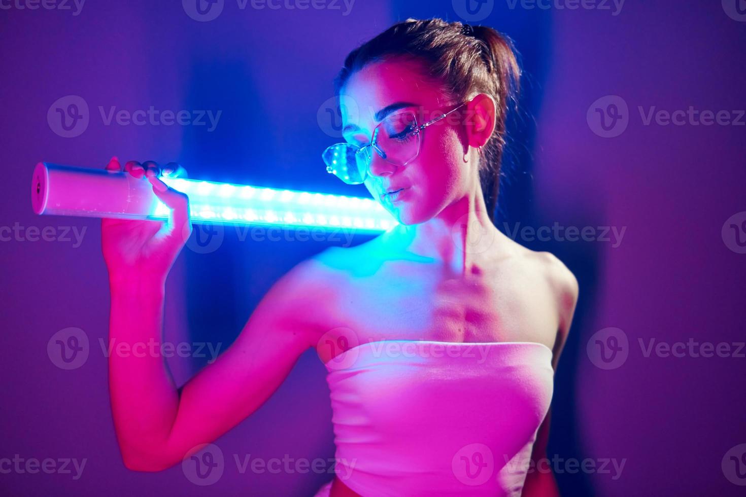 In glasses. Fashionable young woman standing in the studio with neon light photo