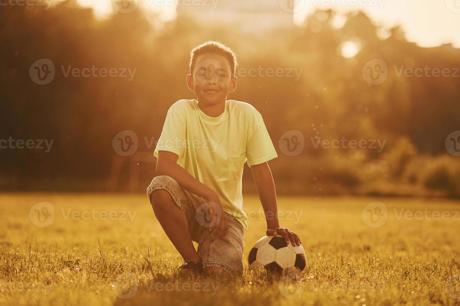Sits with soccer ball. African american kid have fun in the field at summer daytime photo