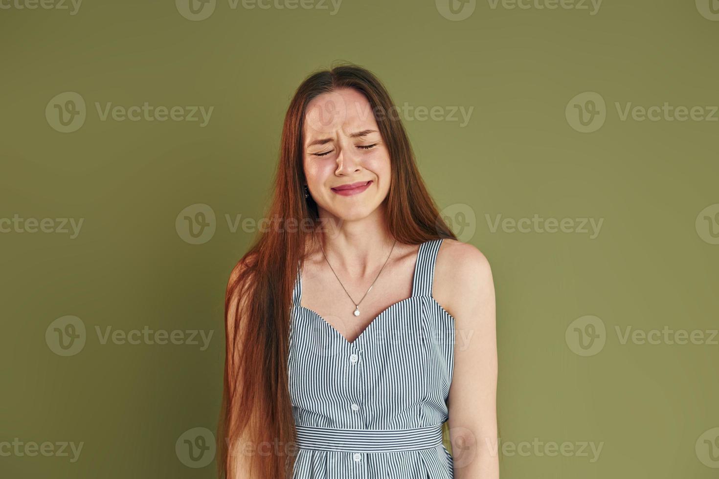 Young woman in casual clothes is crying while standing indoors in the studio photo