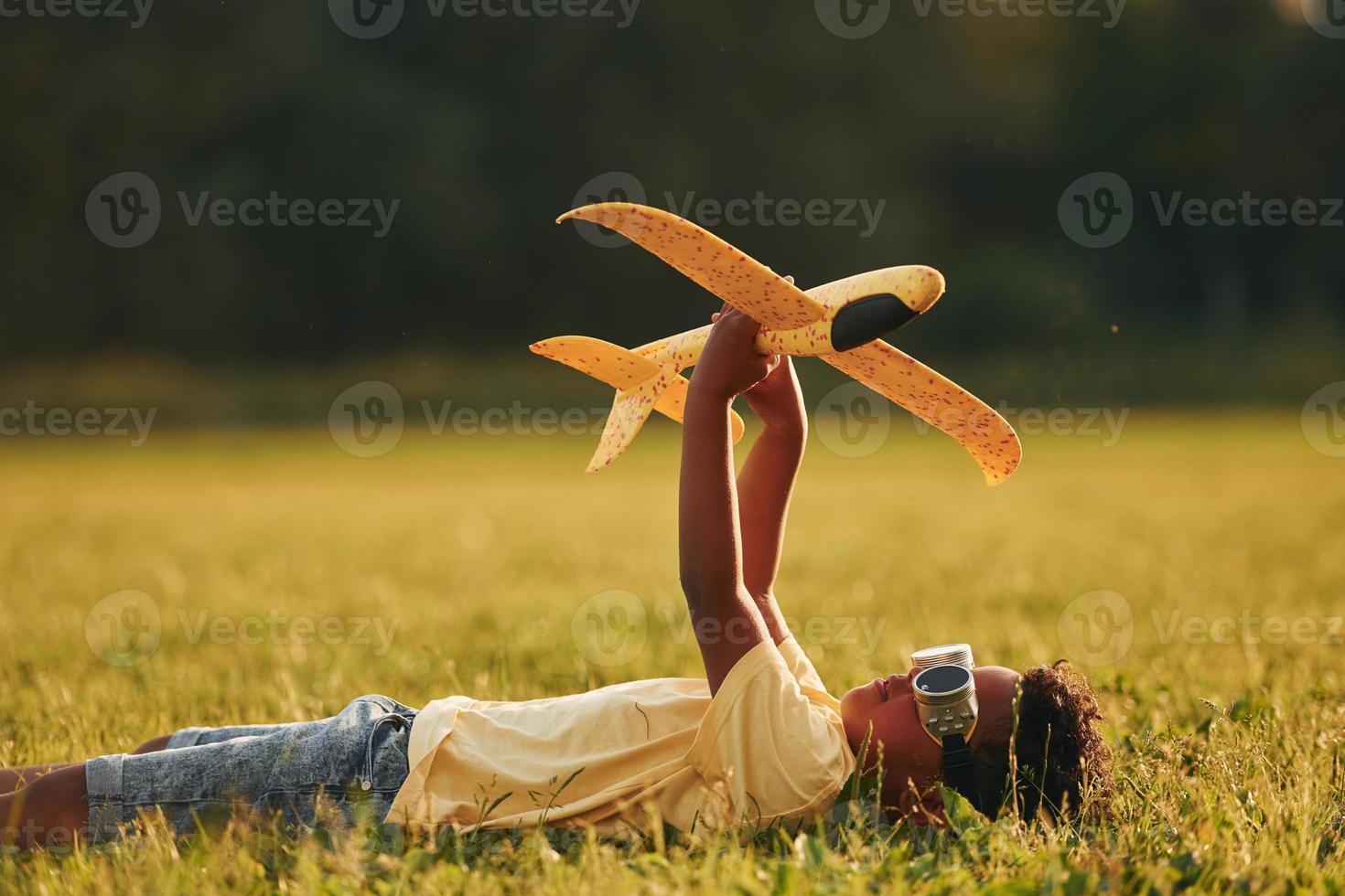 Laying down with toy plane on the grass. African american kid have fun in the field at summer daytime photo