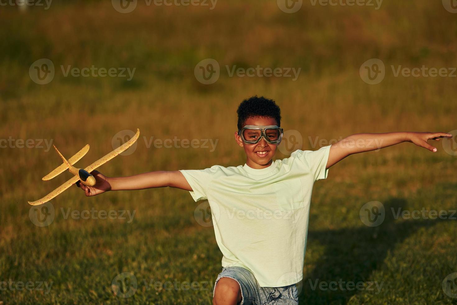 In casual clothes with toy plane. African american kid have fun in the field at summer daytime photo