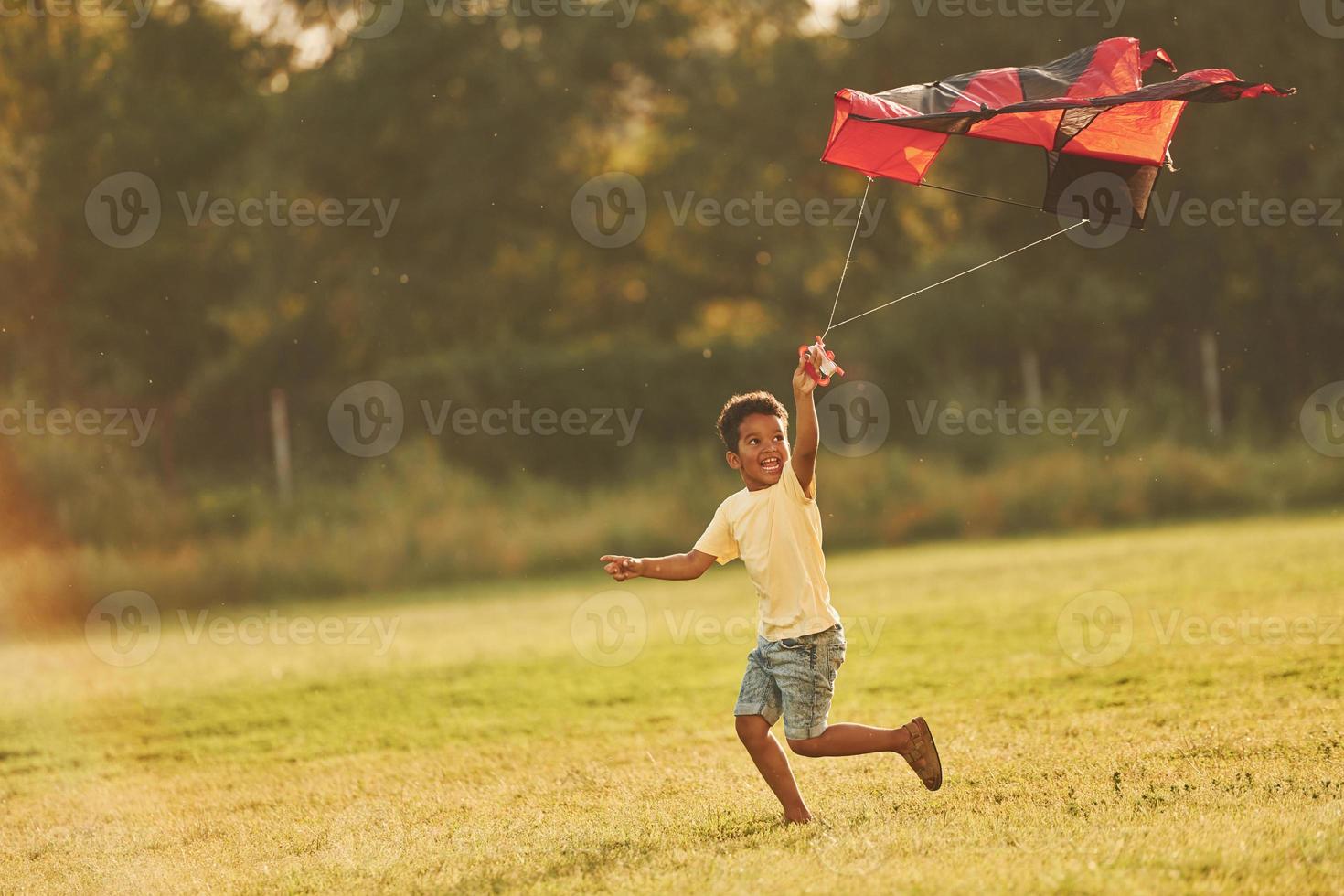 Running with red kite. African american kid have fun in the field at summer daytime photo