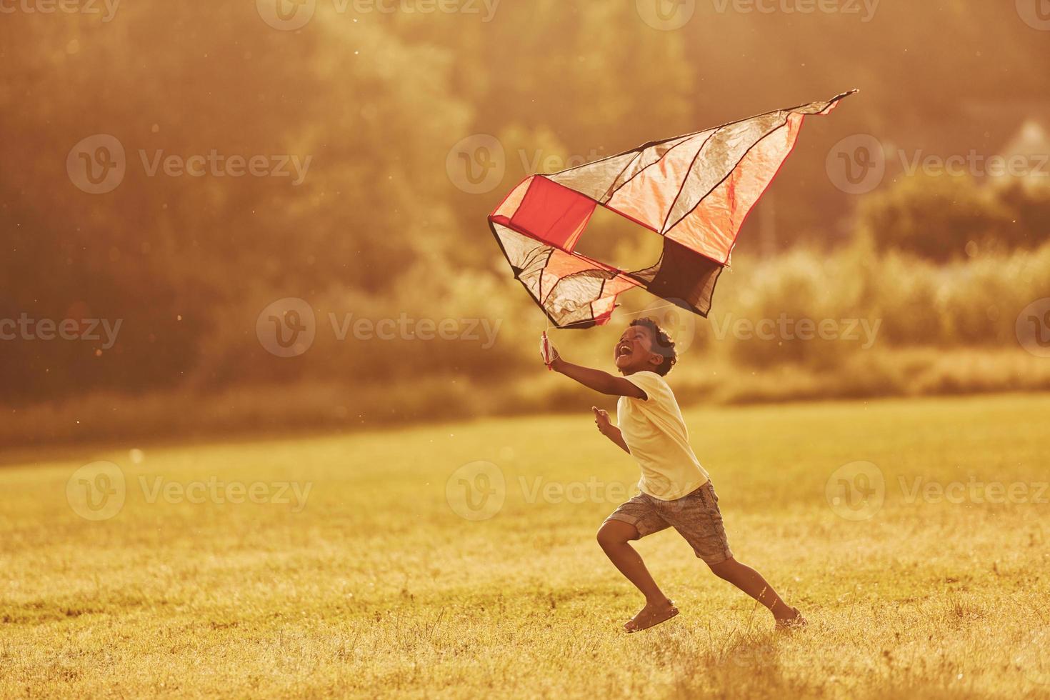 Running with red kite. African american kid have fun in the field at summer daytime photo