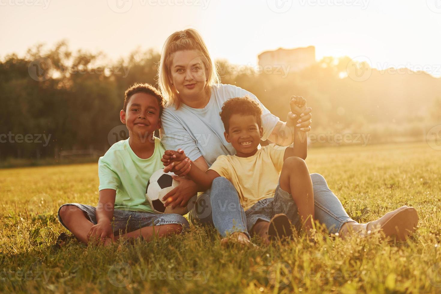 Adult woman sits with two black kids on the field at summer daytime photo