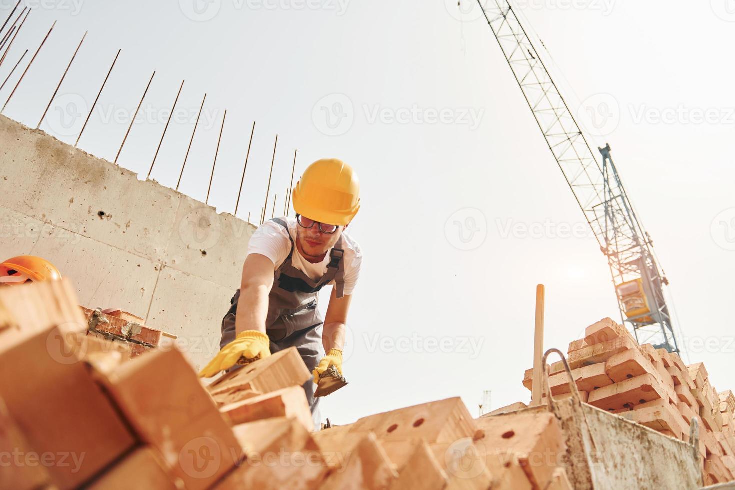 Using bricks. Young construction worker in uniform is busy at the unfinished building photo