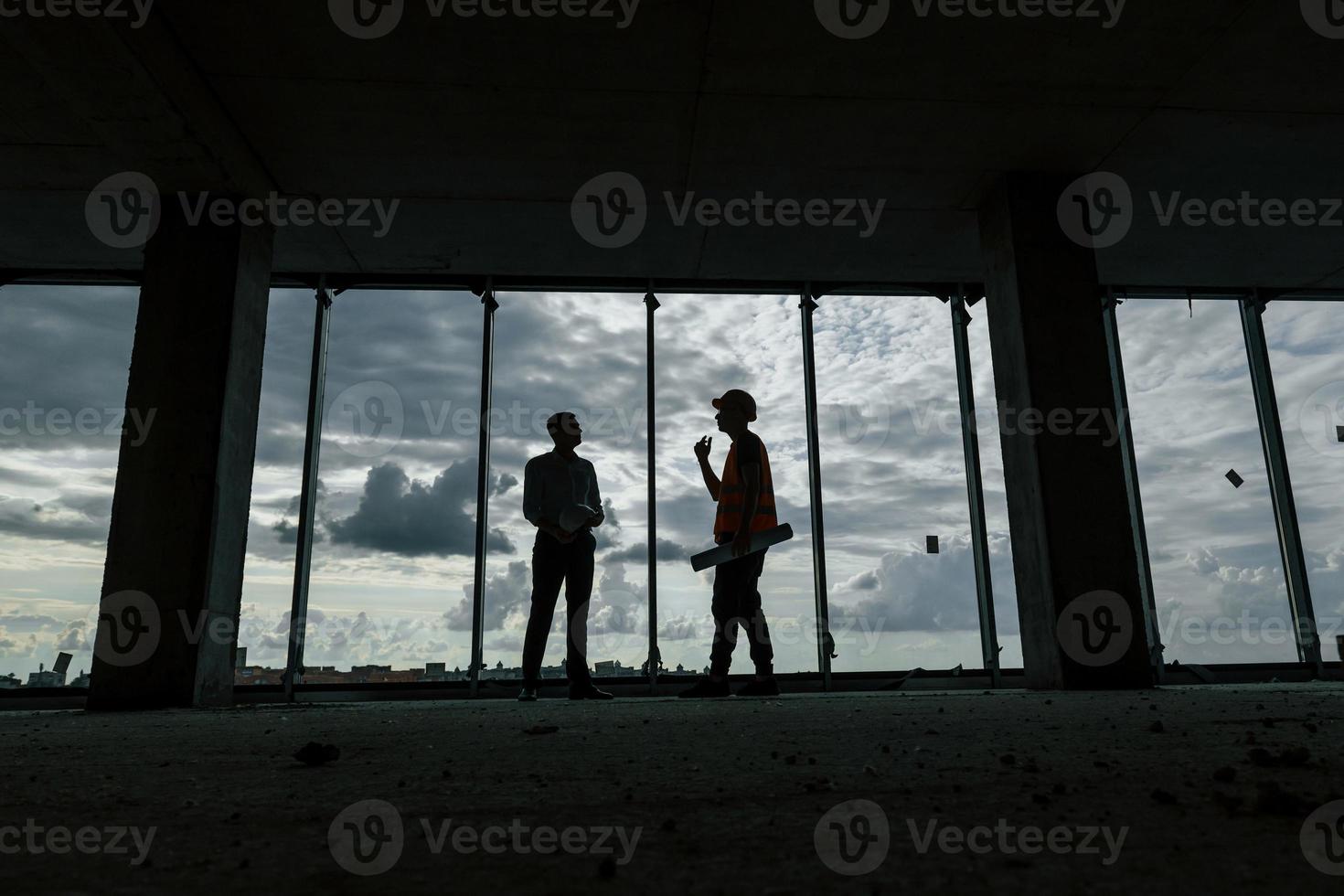 Standing against big window. Man in suit and handyman in orange protective wear is working on construction photo