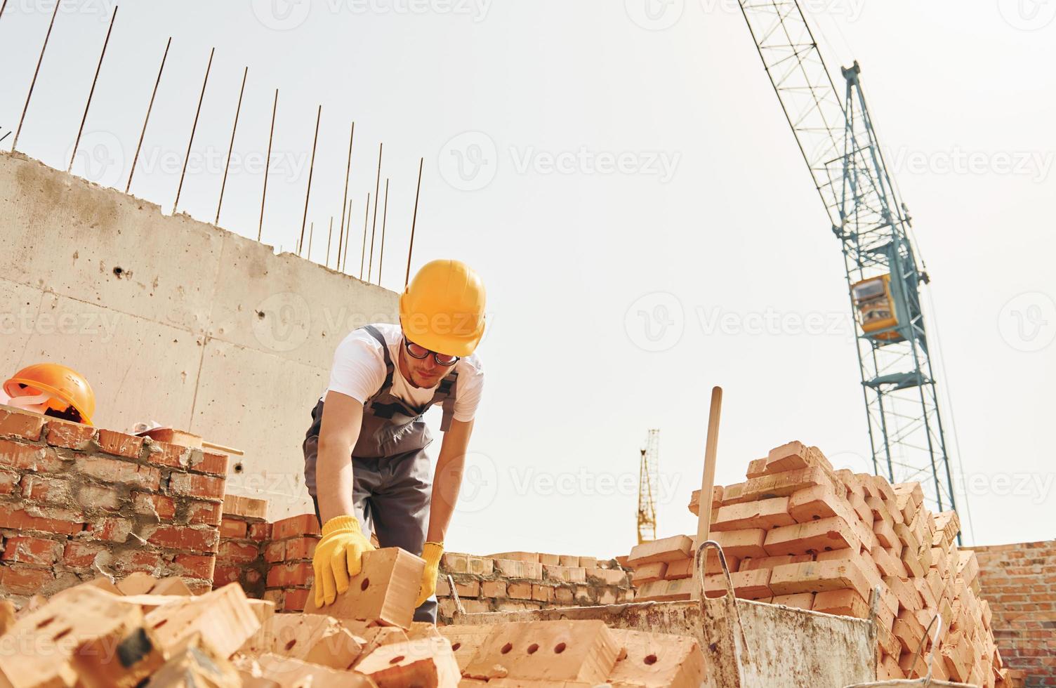 Using bricks. Young construction worker in uniform is busy at the unfinished building photo