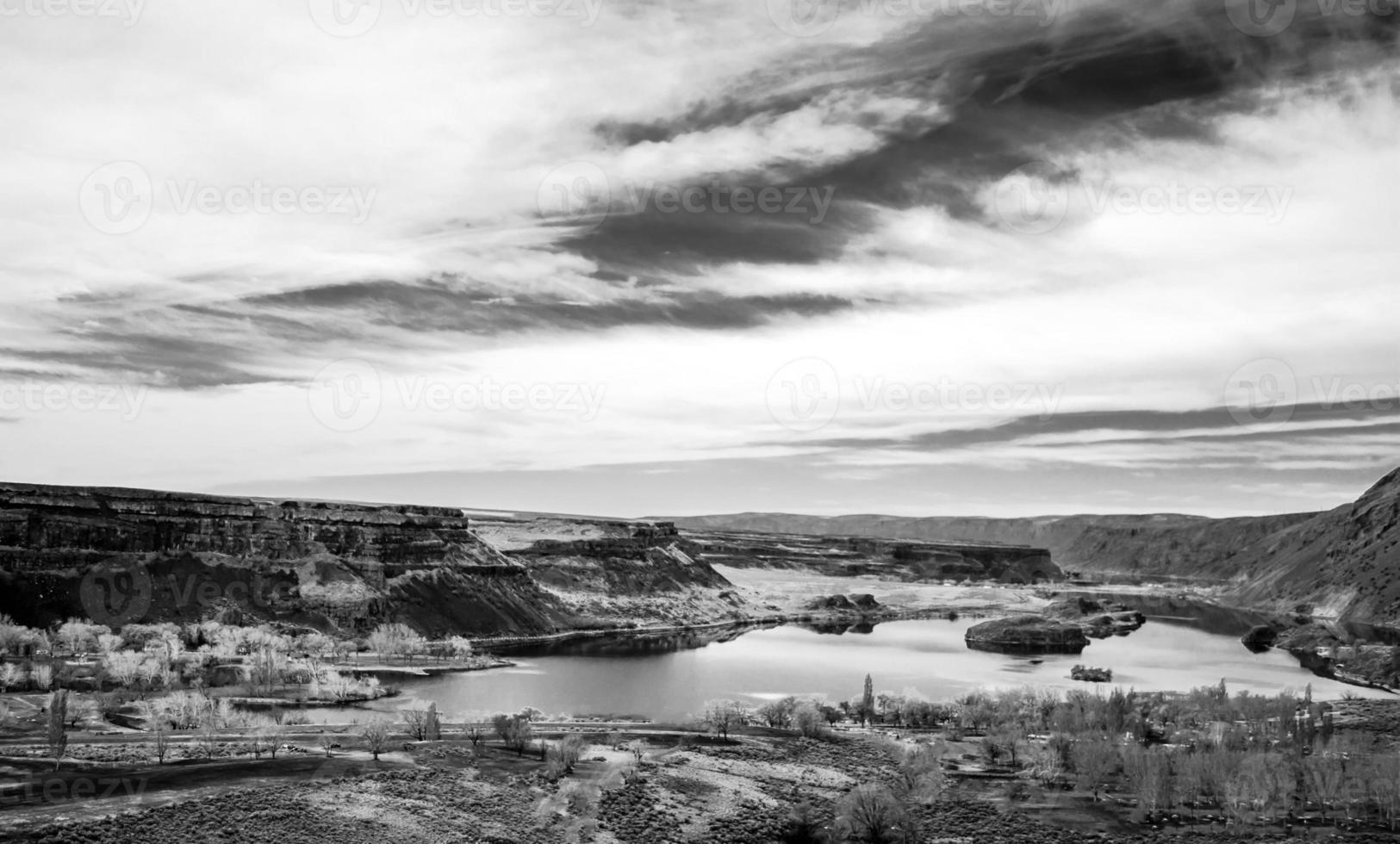 Black and white desert ridge under cloudy sky next to a river photo