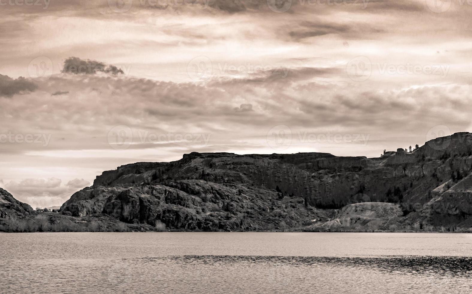 Black and white desert ridge under a cloudy sky next to a river photo
