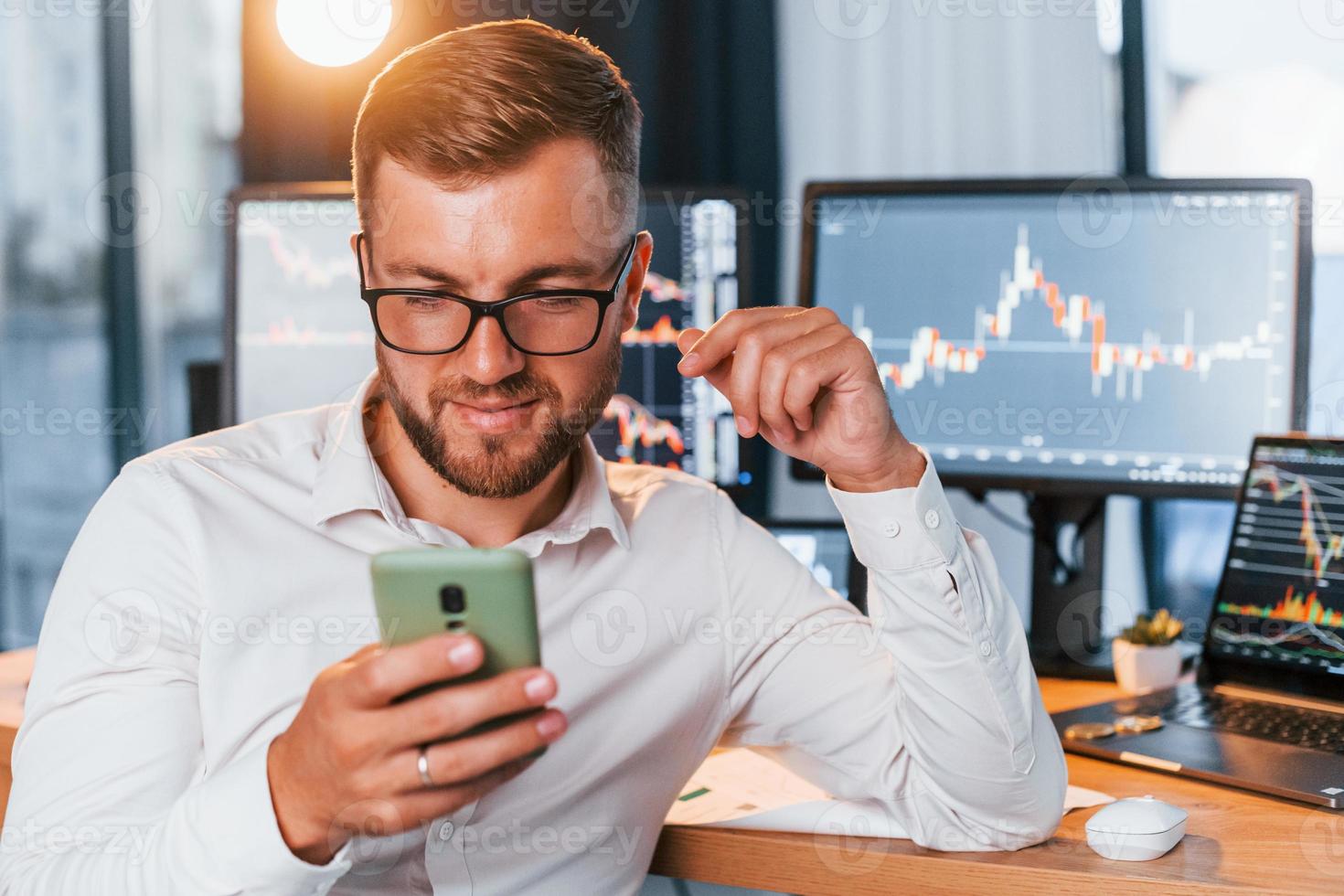 With phone in hands. Young businessman in formal clothes is in office with multiple screens photo