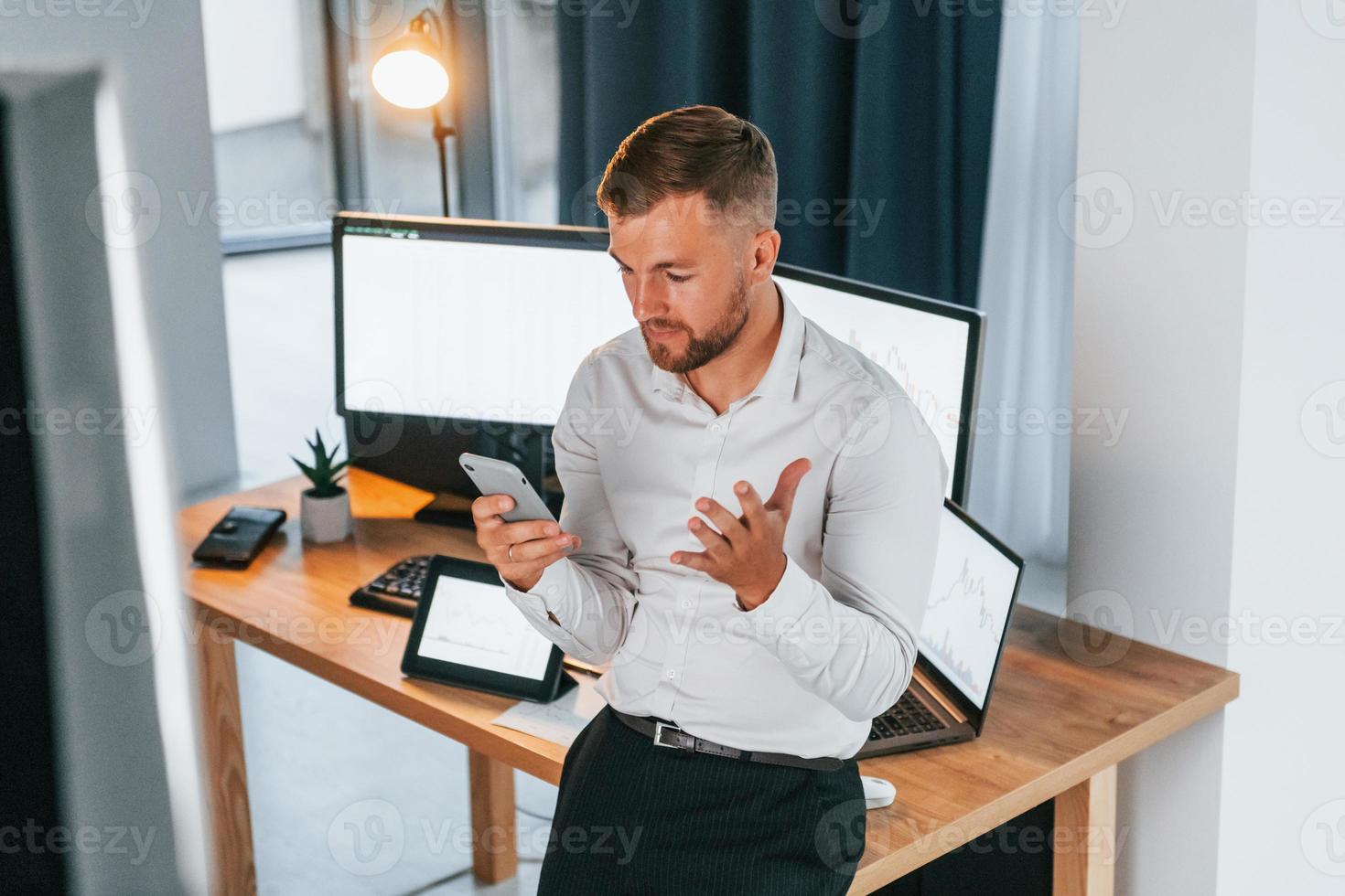 Busy day. Young businessman in formal clothes is in office with multiple screens photo