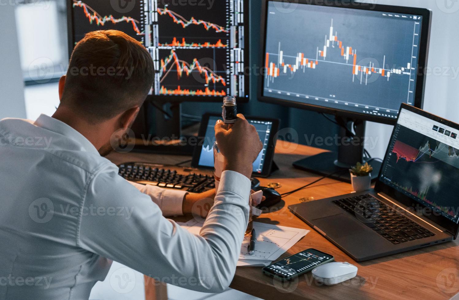 With alcohol. Young businessman in formal clothes is in office with multiple screens photo