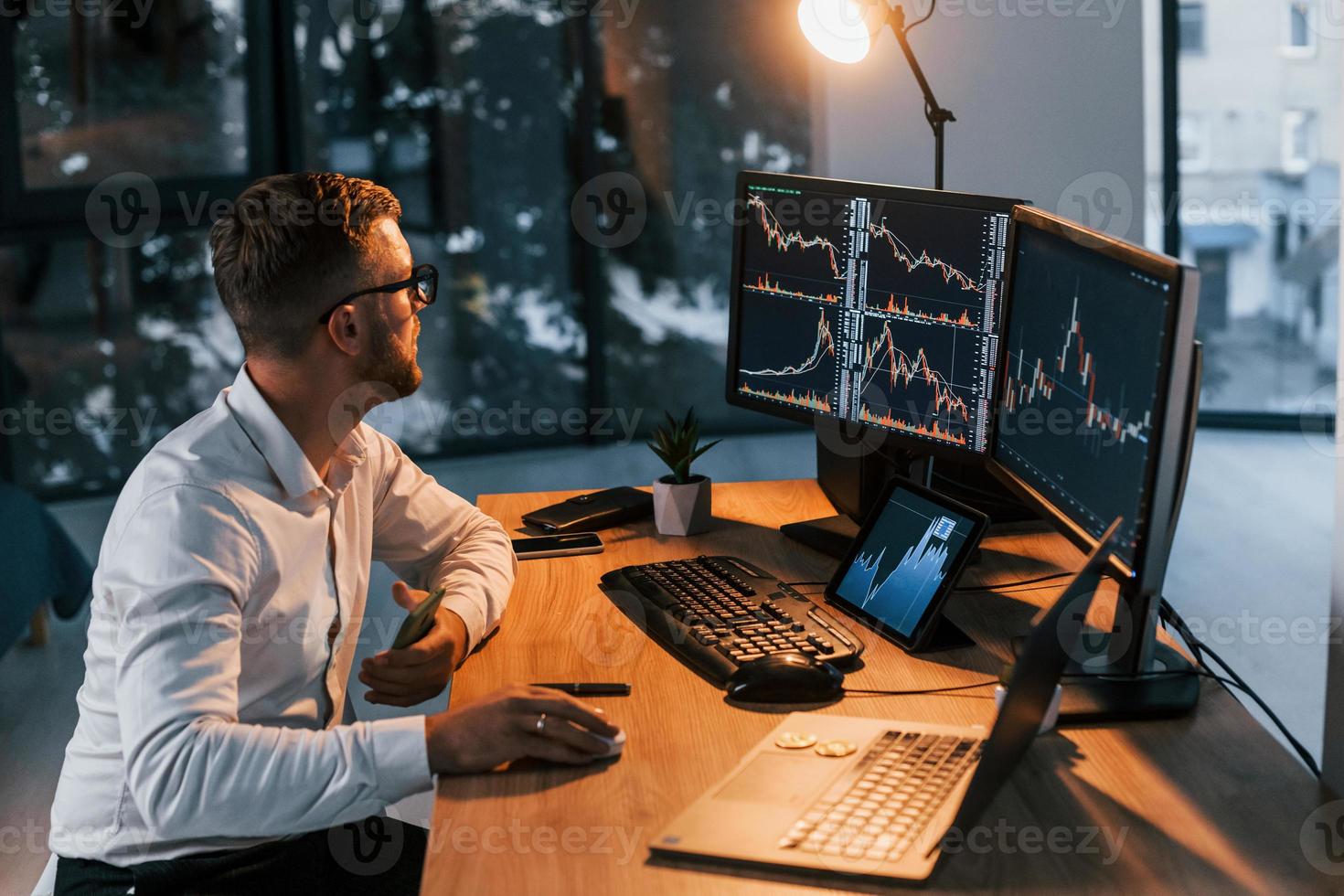 Working at evening time. Young businessman in formal clothes is in office with multiple screens photo
