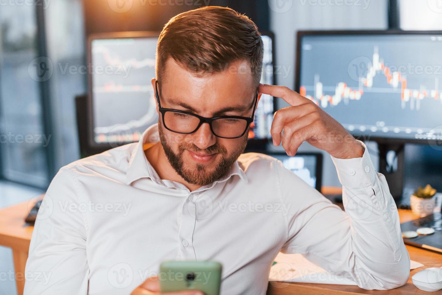 With phone in hands. Young businessman in formal clothes is in office with multiple screens photo