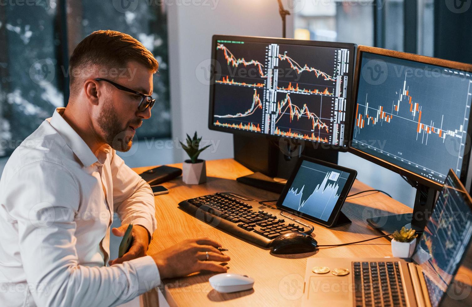 Working at evening time. Young businessman in formal clothes is in office with multiple screens photo