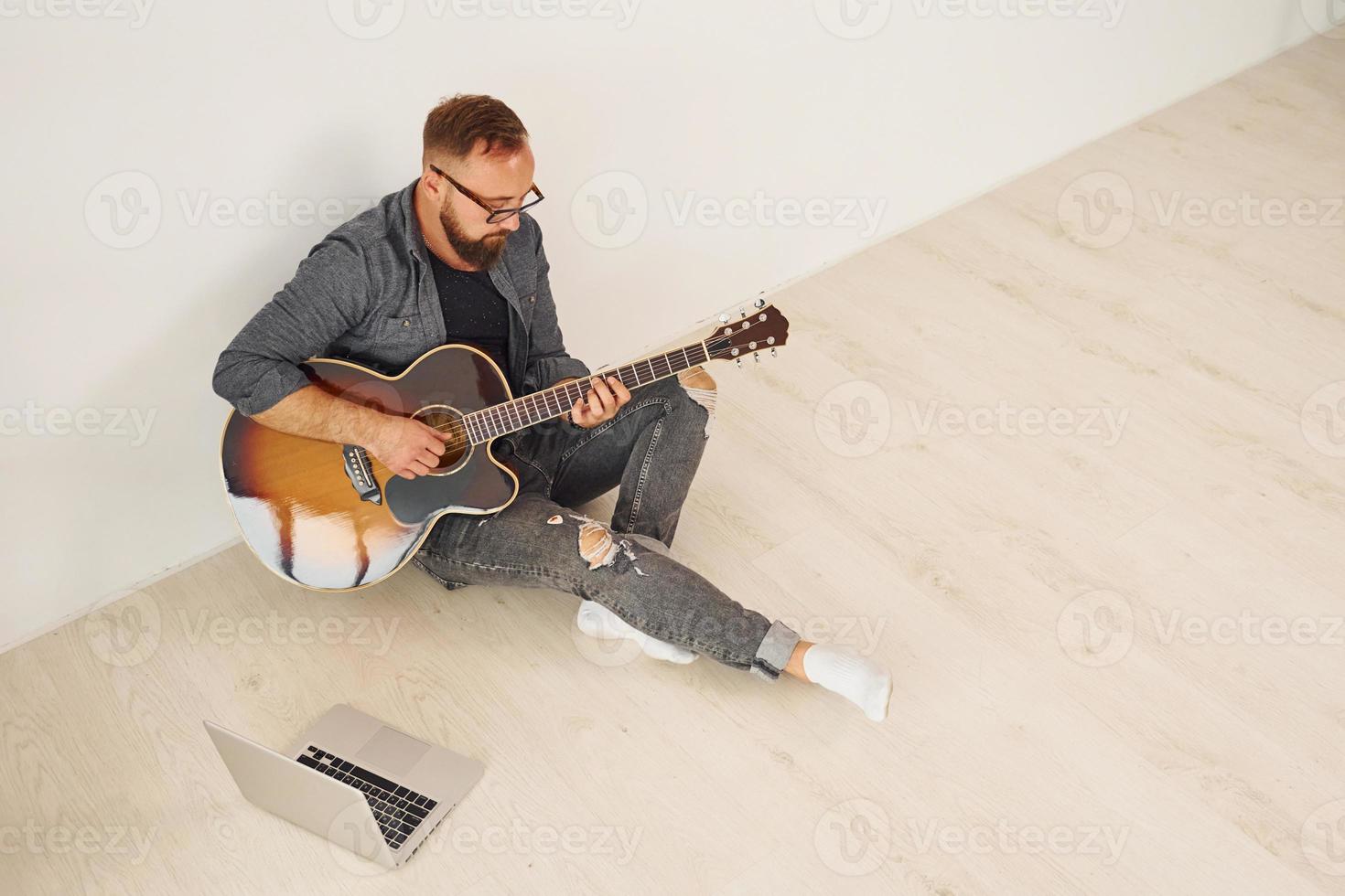 con computadora portátil el hombre con ropa informal y con guitarra acústica está en el interior foto