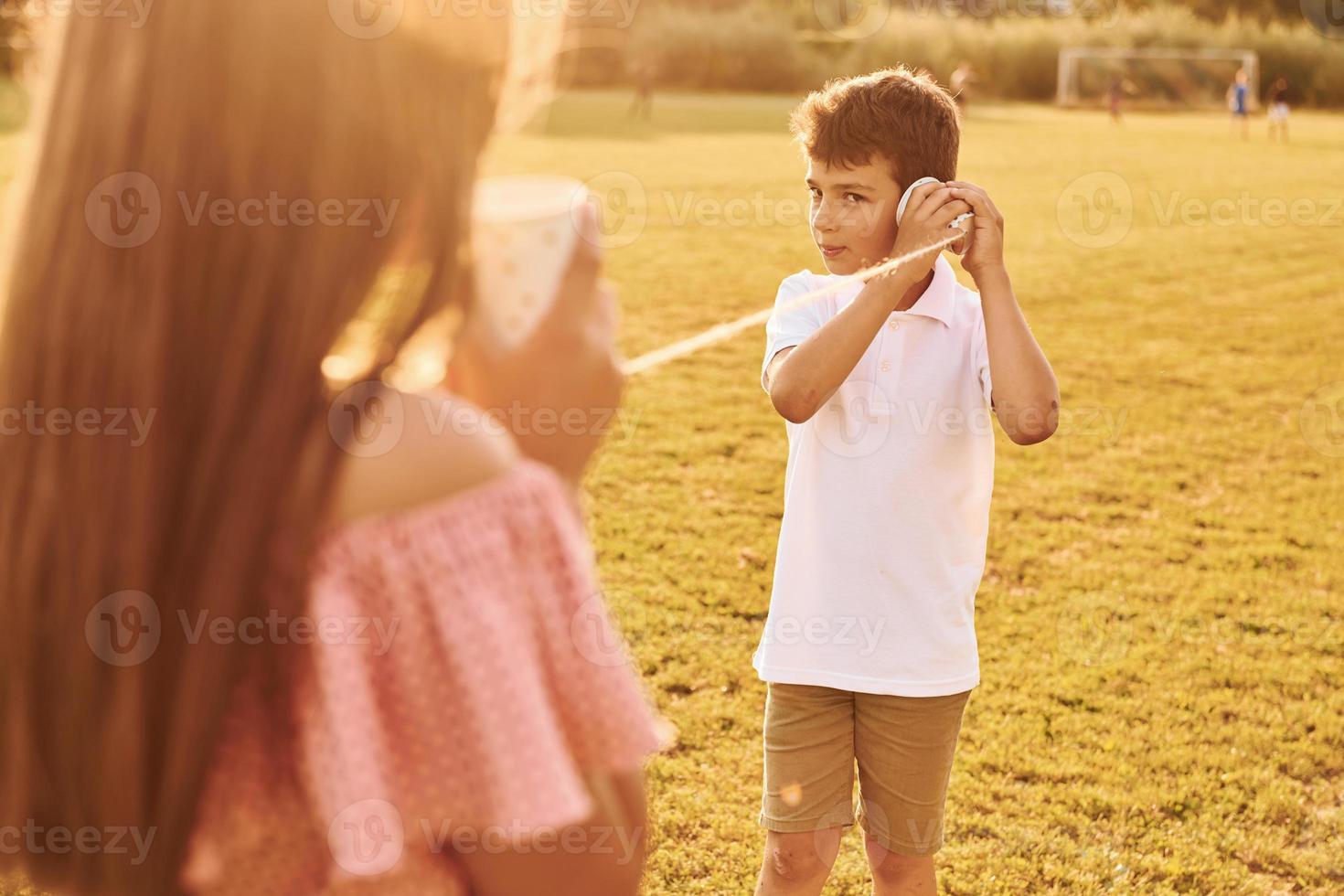 Kids having fun with tin can telephone. Standing outdoors on the sportive field photo
