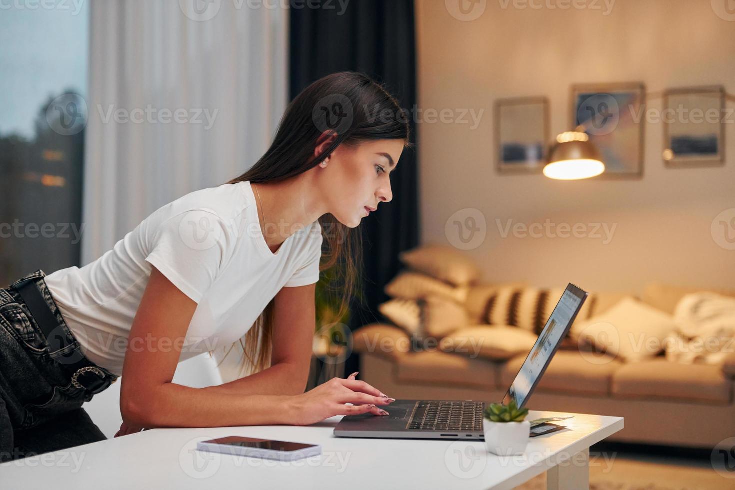 usando una computadora portátil mujer con camisa blanca y jeans negros está de pie en el interior de su casa foto