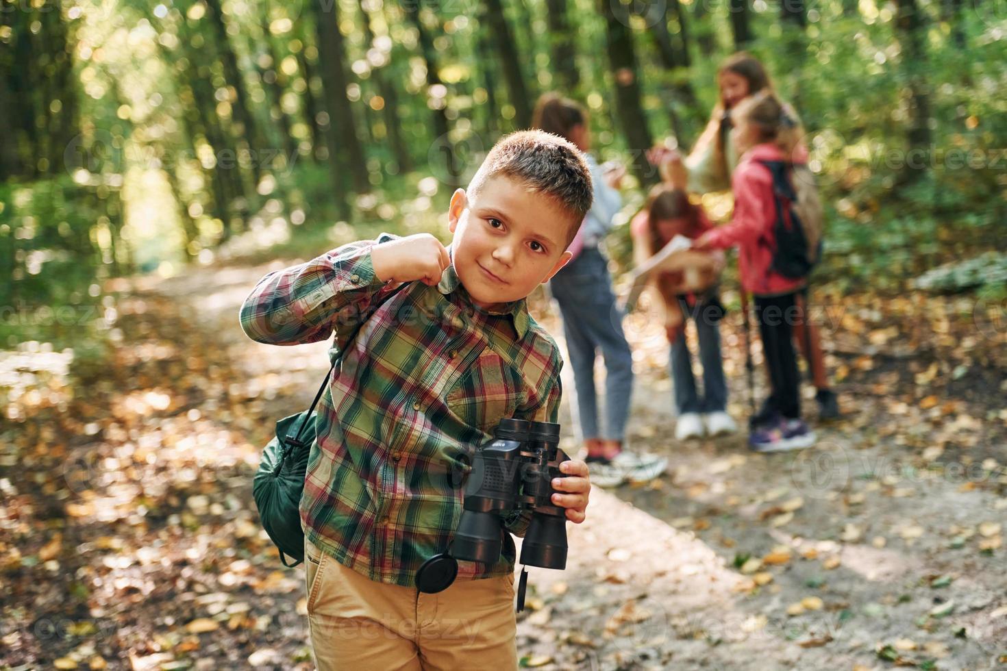 Boy with binoculars standing in front of his friends. Kids in green forest at summer daytime together photo