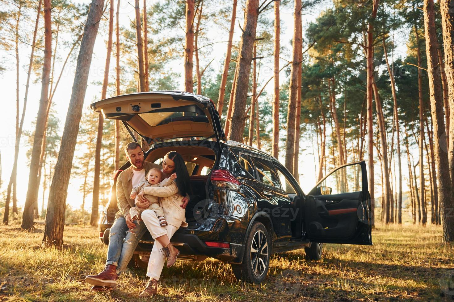 With black colored automobile. Happy family of father, mother and little daughter is in the forest photo