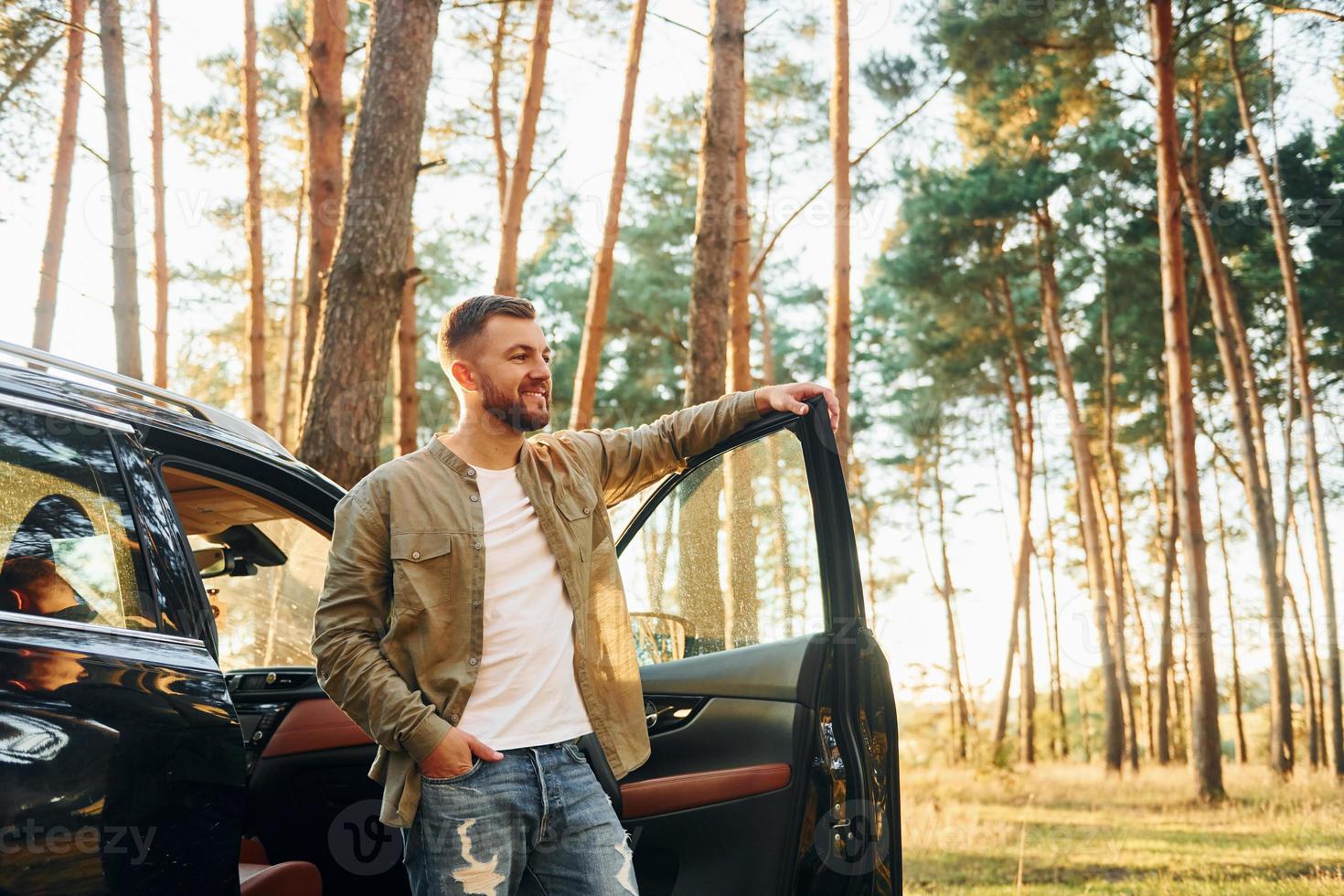 With his car. Man in jeans is outdoors in the forest with his black colored automobile photo