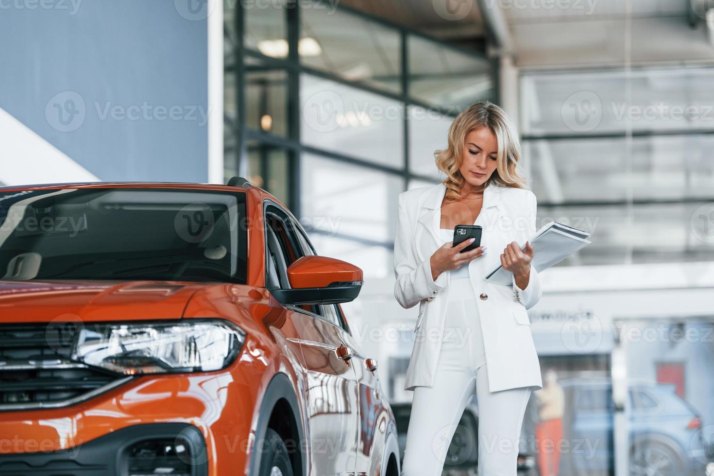 Holding phone. Woman in formal clothes is indoors in the autosalon photo