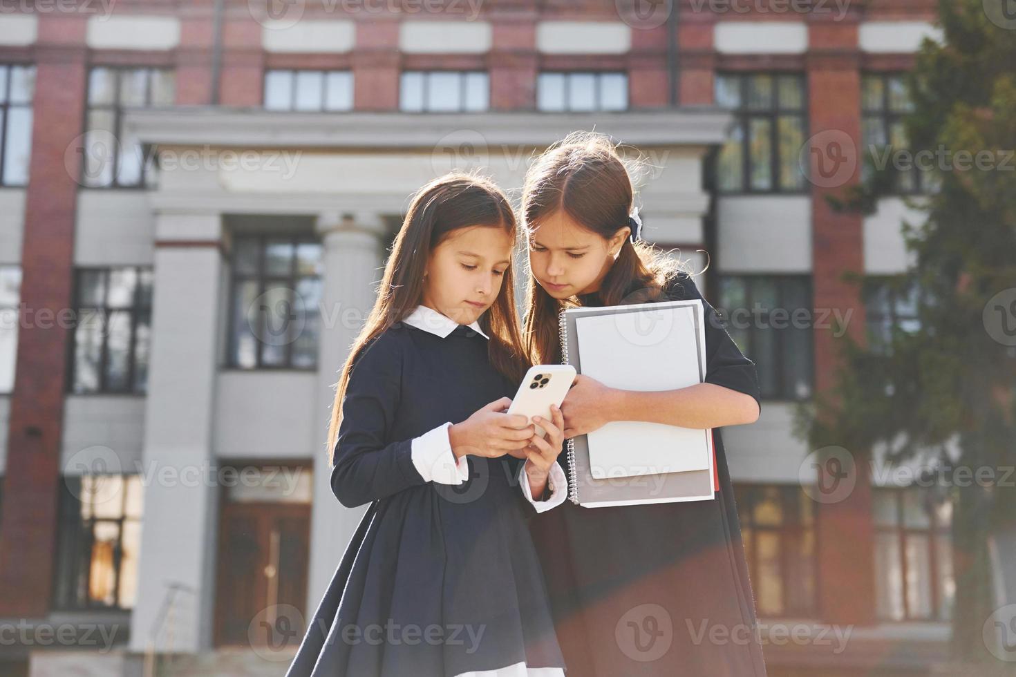 Holding books. Two schoolgirls is outside together near school building photo