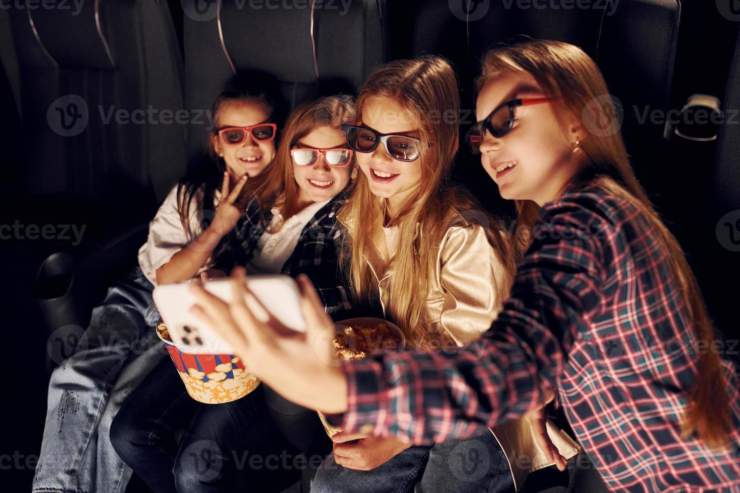 Holding phone in hand. Group of kids sitting in cinema and watching movie together photo