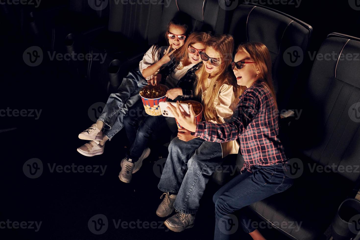 Holding phone in hand. Group of kids sitting in cinema and watching movie together photo