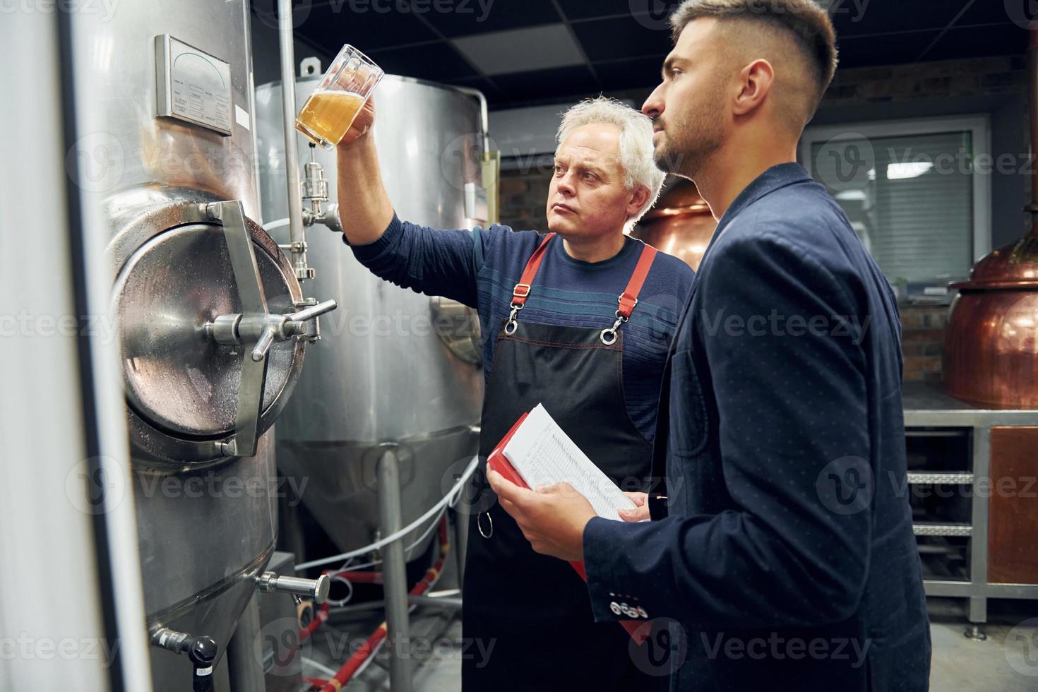 Holding glass. Senior man and young worker with document is standing in the storage beer room photo