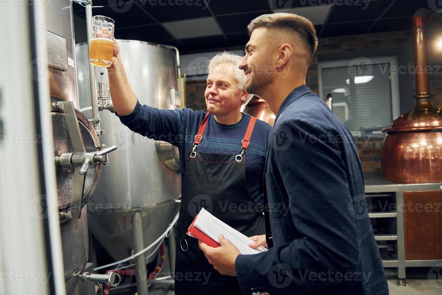 Holding glass. Senior man and young worker with document is standing in the storage beer room photo