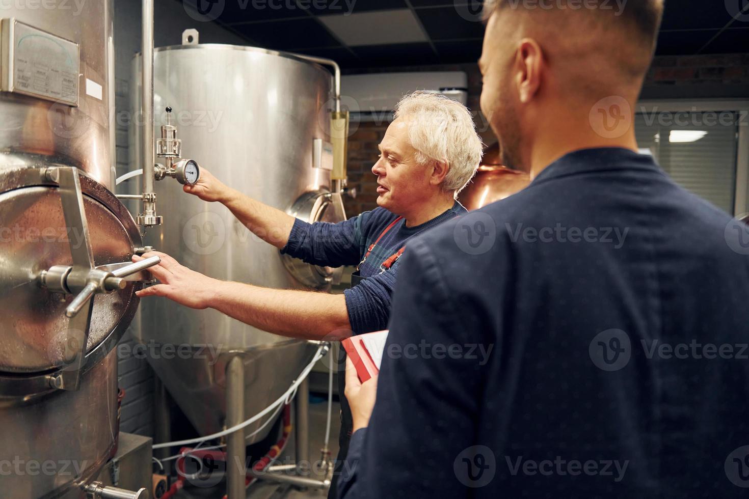 un anciano y un joven trabajador con documento están parados en la sala de cerveza de almacenamiento foto