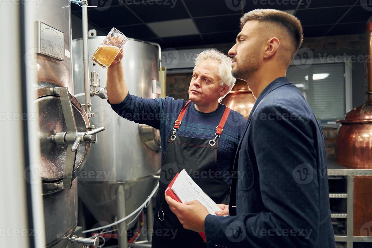 Holding glass. Senior man and young worker with document is standing in the storage beer room photo