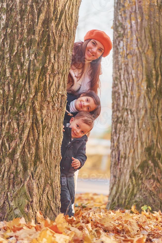 escondiéndose detrás de un árbol. madre con su hijo pequeño y su hija se divierten en el parque de otoño foto